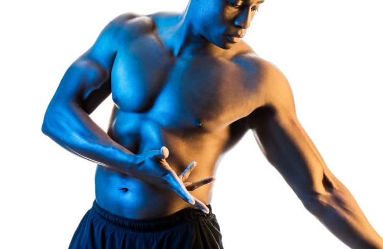 Man with no shirt and black shorts facing the camera with a blue glow to his skin, head tilted down and arms flexed