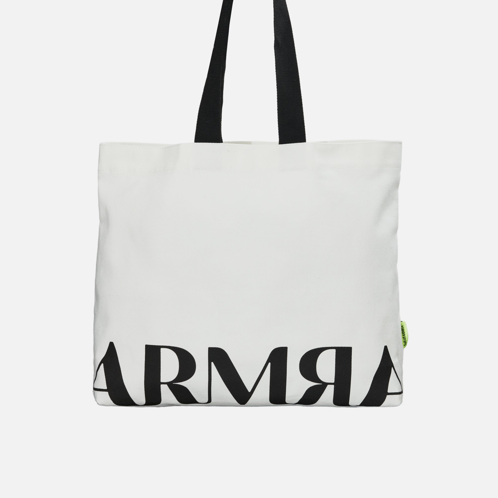 Front of white tote bag with black handles hanging with black text “ARMRA”