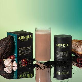 Tall clear glass Chocolate ARMRA Colostrum™ Performance Revival surrounded by a jar of Performance Revival, Performance Revival packaging and cacao fruit. 