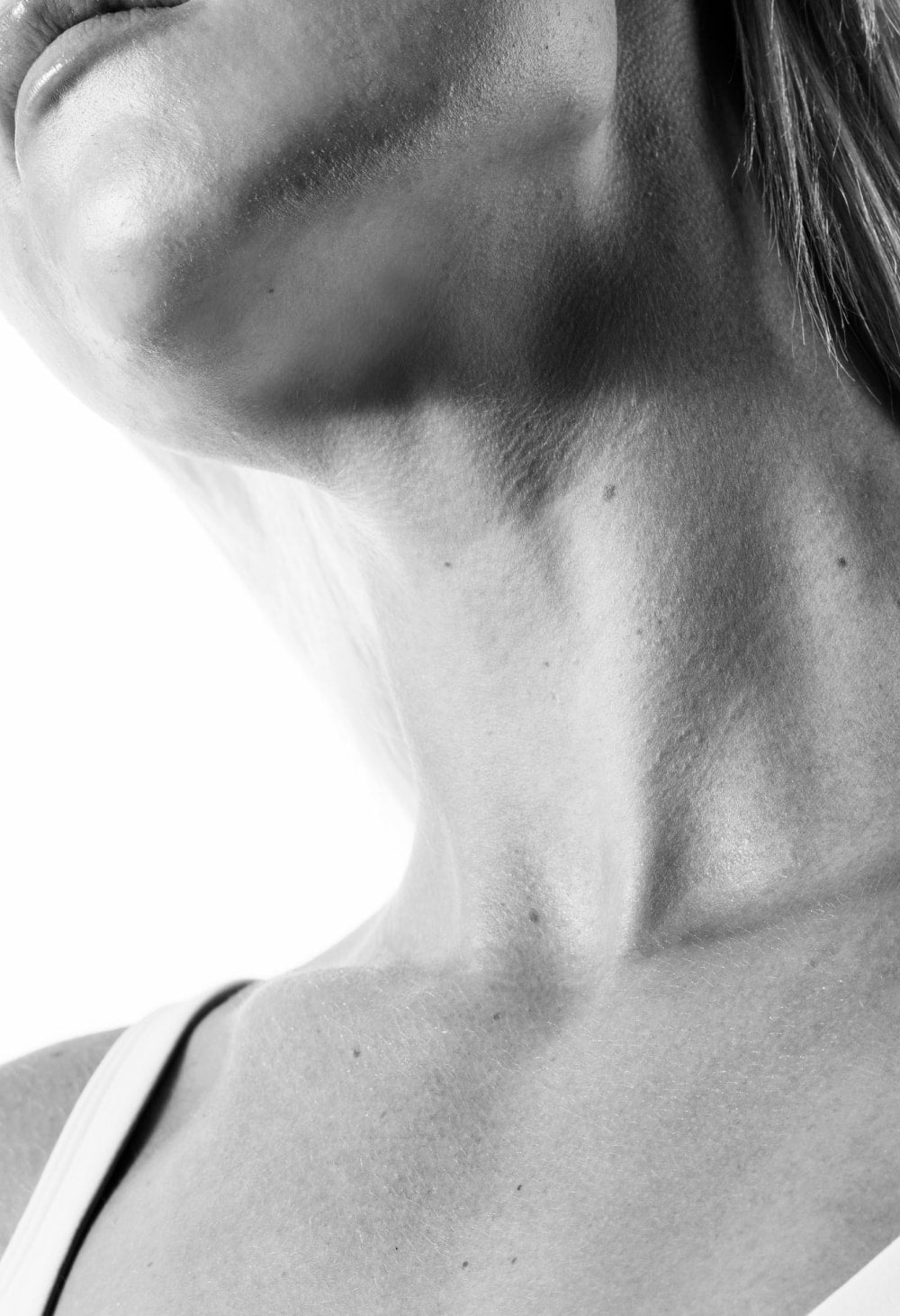 A black and white cropped image showing a womans chest, neck, chin tilted up, and lips wearing a white tank top on a white background