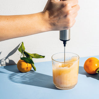 Person holding stainless steel whirl, stirring orange powder into a clear glass on a light blue table top and white background with oranges behind