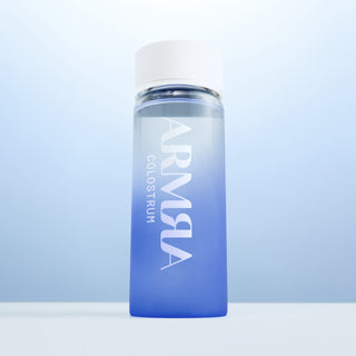 Cream Lid on a Glass Water Bottle with blue gradient and “ARMRA Colostrum “Logo on a tabletop with blue background