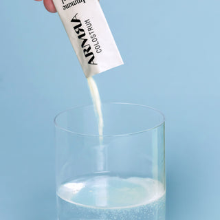 Hand holding a white Stick Pack pouring out colostrum into a clear glass of water on a blue background